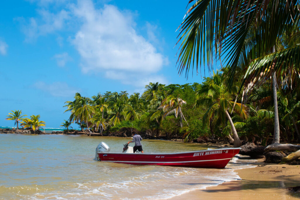 A person stands in a red motorboat named “Siete Hermanos” near the shore of a tropical beach, surrounded by lush palm trees and clear blue sky—an ideal moment on a 10-Day Nicaragua Itinerary.