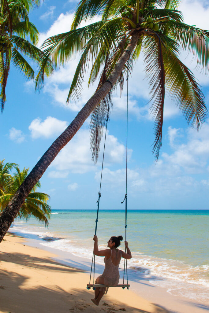 A woman in a swimsuit sits on a swing hanging from a leaning palm tree on a sandy tropical beach—a dreamy moment you might find on your 10-Day Nicaragua Itinerary, with turquoise ocean water and blue sky in the background.
