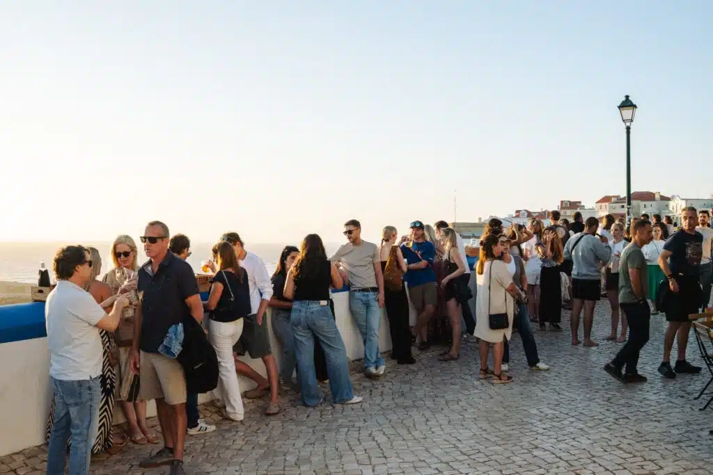 A large group of people stands and socializes on a sunlit cobblestone terrace overlooking the sea, near some of the Best Restaurants In Ericeira, with houses and a lamppost in the background under a clear sky.