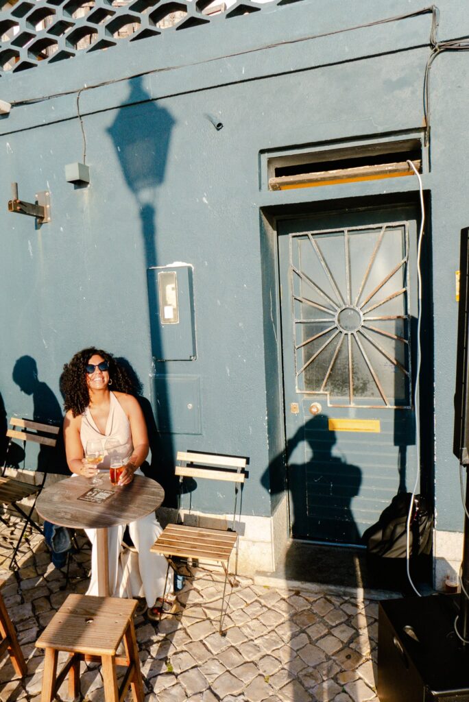A woman with curly hair and sunglasses sits at a small outdoor table with drinks, beside a blue wall and door—enjoying sunlight, perhaps outside one of the best restaurants in Ericeira. Shadows of a streetlamp and another person are cast nearby.