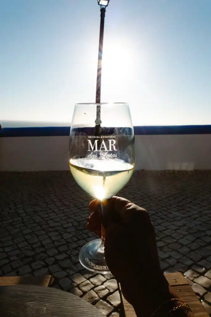 A hand holds a wine glass with the word "MAR" printed on it, raised against a sunlit sky—perfect for savoring the atmosphere near some of the Best Restaurants In Ericeira, with a cobblestone patio and blue and white wall in the background.