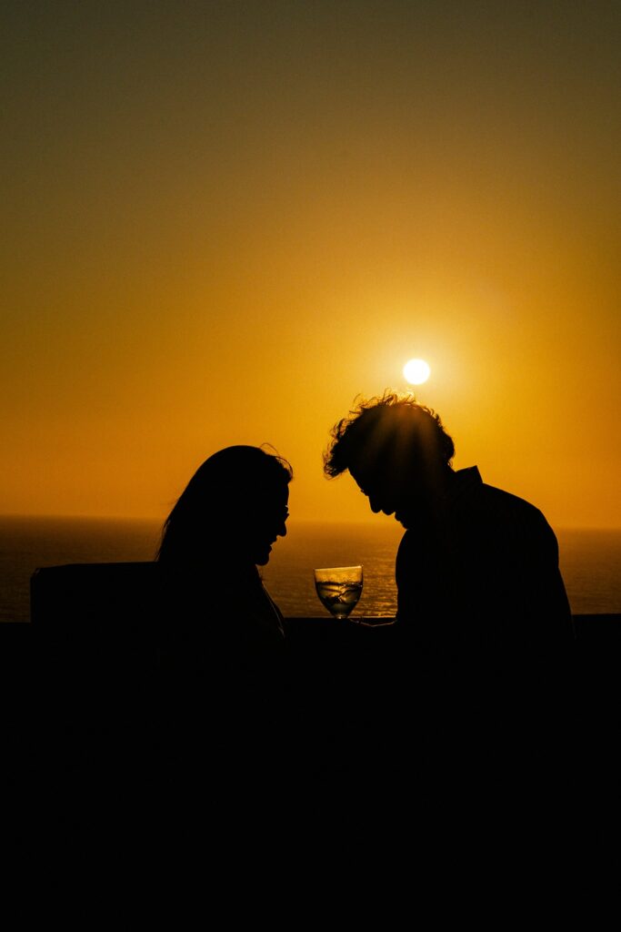 Silhouette of two people facing each other at sunset, a wine glass between them, with the sun setting over the ocean—capturing the romantic ambiance found at some of the Best Restaurants In Ericeira.