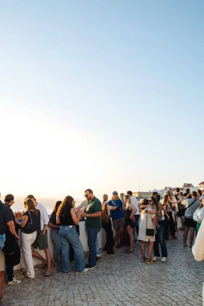 A large group gathers outdoors on a cobblestone terrace, socializing and enjoying drinks under a clear blue sky at sunset, with the ocean in the background—an experience typical of the best restaurants in Ericeira.