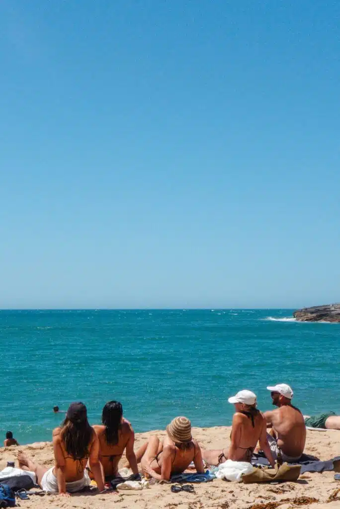Five people in swimwear sit on a rocky beach facing the turquoise ocean, chatting and relaxing. With swimmers in the background, it's the perfect setting to discuss plans for visiting the Best Restaurants In Ericeira nearby.