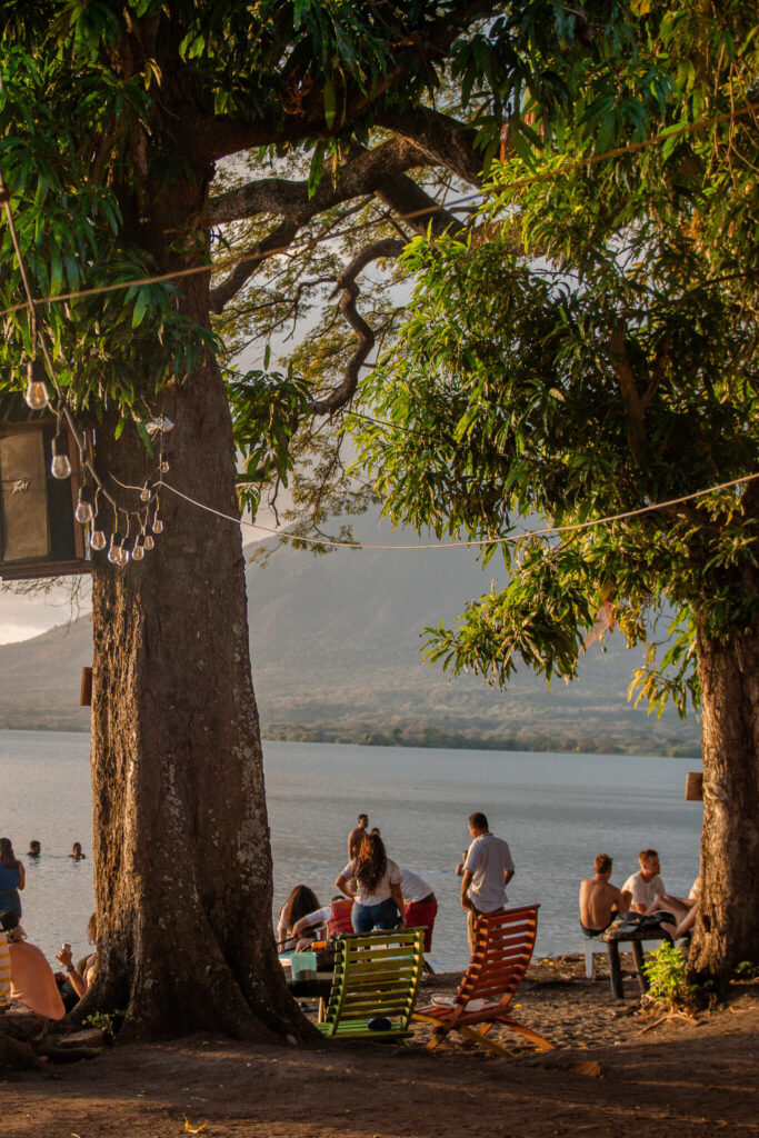 People relax by a lakeshore under large trees, with string lights overhead—an ideal scene to include in your 10-Day Nicaragua Itinerary. Some lounge in colorful chairs, others stand or swim as mountains and a partly cloudy sky fill the background.