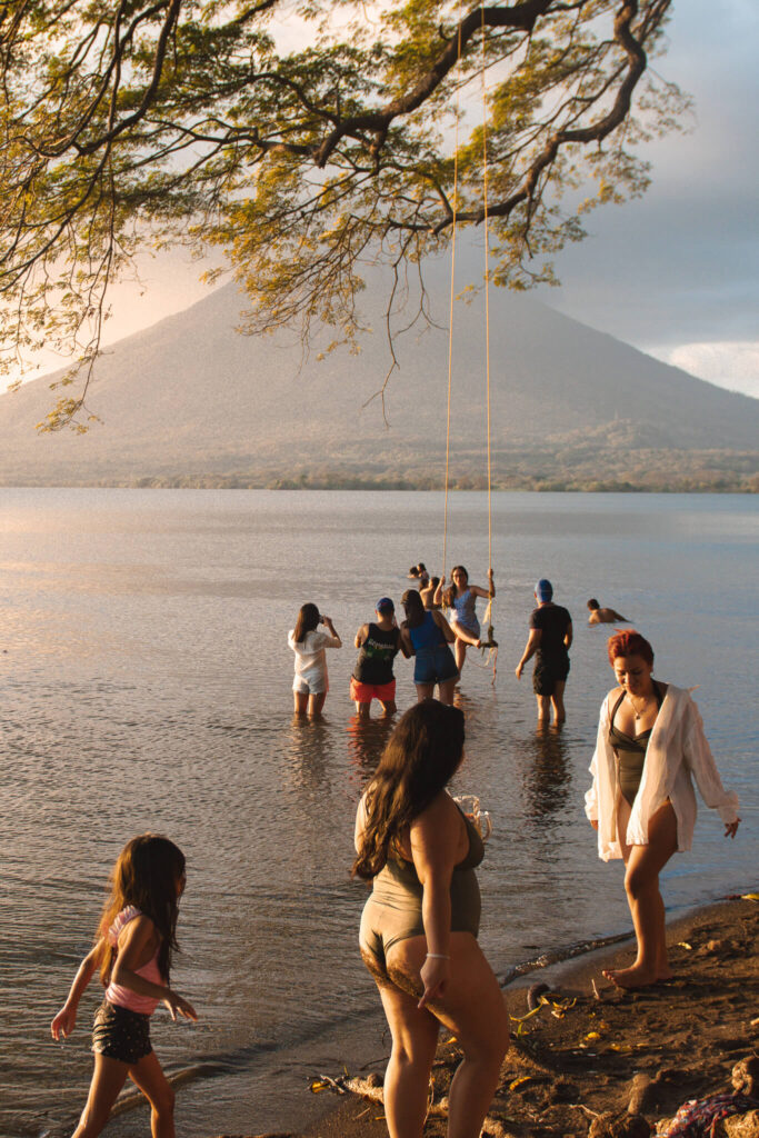 A group of people enjoy a lakeside scene during their 10-Day Nicaragua Itinerary, some standing in shallow water and others using a rope swing. Trees frame the image, with a mountain rising in the background under a cloudy sky.