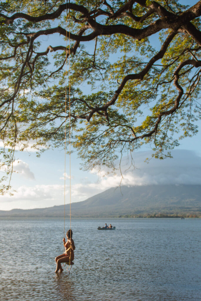 A person enjoys a rope swing hanging from a large tree over calm water, with distant mountains and a small boat in the background—an idyllic scene perfect for your 10-Day Nicaragua Itinerary under a partly cloudy sky.
