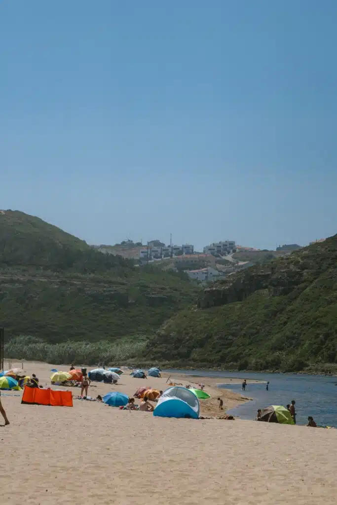 People relax under colorful beach tents on a sandy shore in Portugal, bordered by green hills and calm waters. Distant buildings atop the hills hint at some of the Best Things To Do In Ericeira under a clear blue sky.