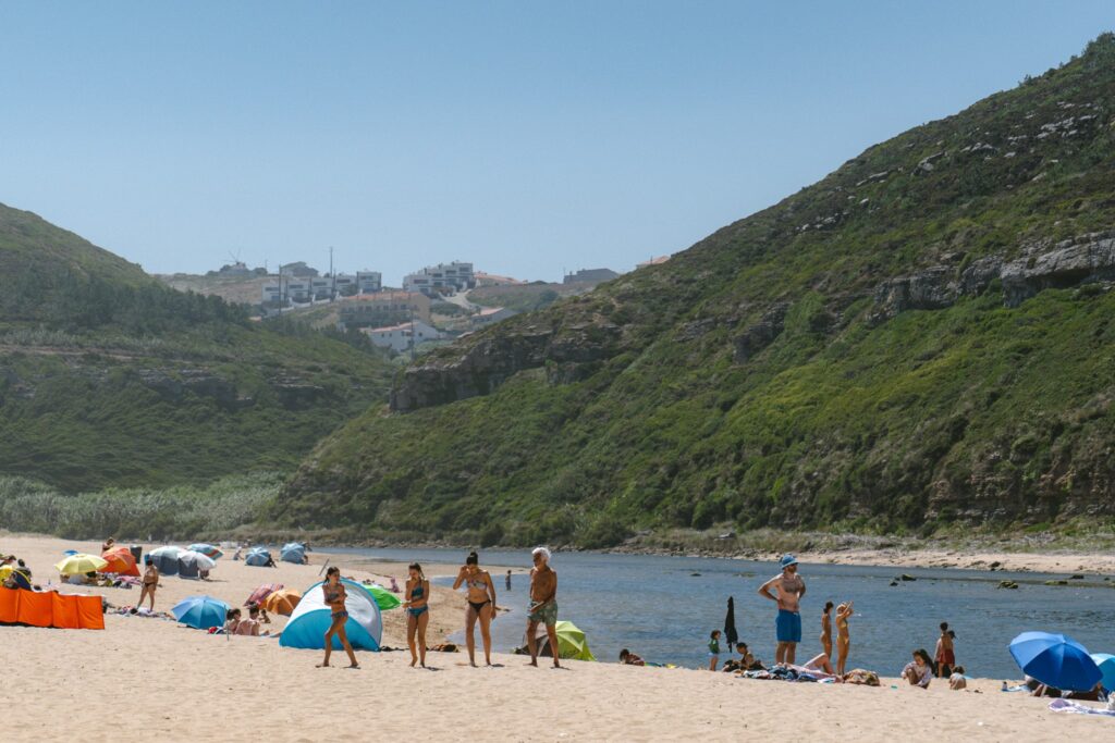 People relax on a sandy river beach in Portugal, with colorful umbrellas and towels. Green hills rise in the background, and distant buildings dot the hill under a clear blue skyโa scene among the Best Things To Do In Ericeira.