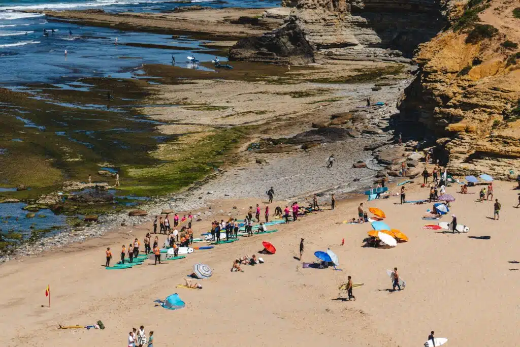 A sandy beach with colorful umbrellas, people sunbathing, and groups taking surfing lessons near rocky cliffs and shallow tide pools under a clear sky—one of the Best Things To Do In Ericeira, Portugal.
