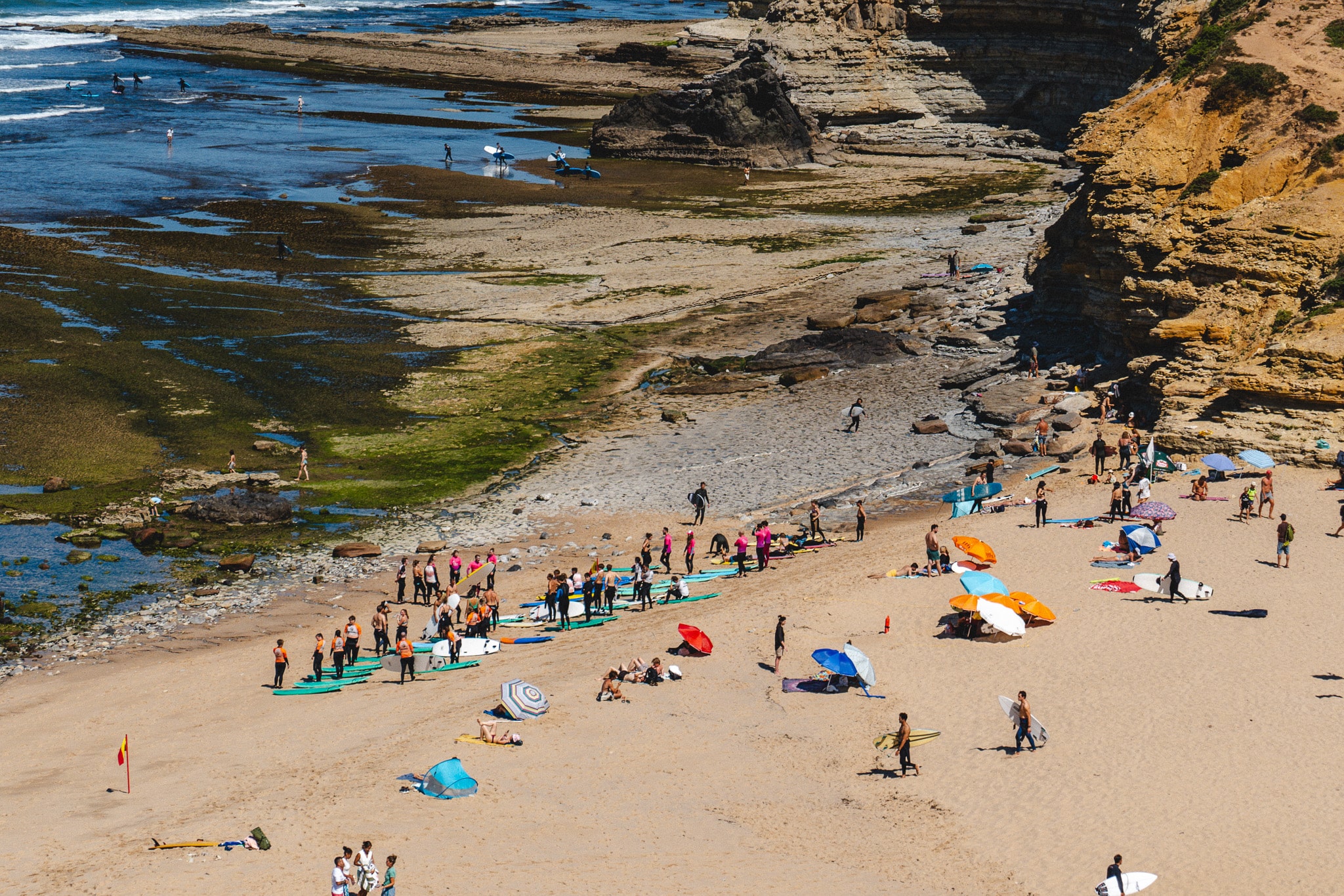 A sandy beach with colorful umbrellas, people sunbathing, and groups taking surfing lessons near rocky cliffs and shallow tide pools under a clear sky—one of the Best Things To Do In Ericeira, Portugal.