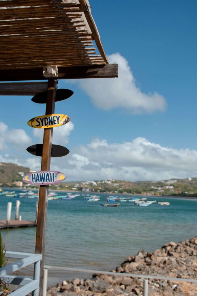 A wooden structure with surfboard-shaped signs reading "SYDNEY" and "HAWAII" stands by a rocky shoreline, evoking the adventure of a 10-Day Nicaragua Itinerary, with boats drifting on calm blue water under a sunny sky.