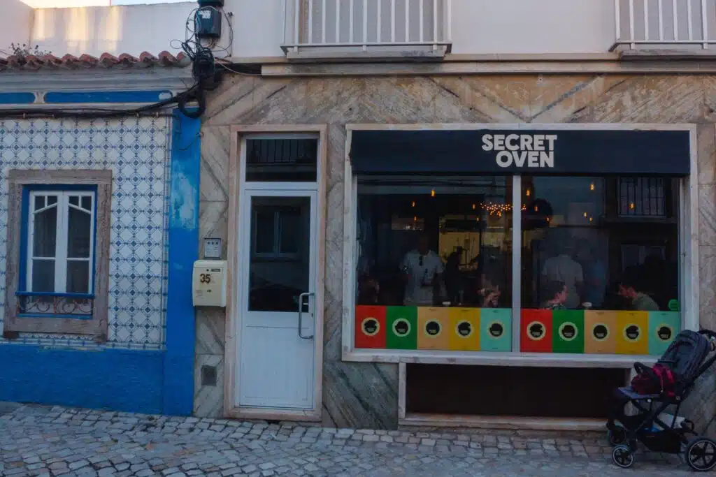 A street view shows a tiled building and the entrance to "Secret Oven," one of the best restaurants in Ericeira, with people inside and a stroller parked outside on the cobblestone sidewalk.