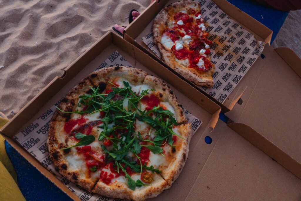 Two open pizza boxes on a sandy surface; one holds a whole mozzarella, tomato, and fresh arugula pizza, while the other has pepperoni slices—just what you’d expect from the Best Restaurants In Ericeira.
