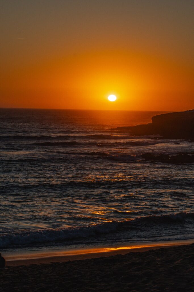 Sun setting over the ocean near Ericeira, casting orange and yellow hues across the sky and water. Gentle waves roll toward the sandy shore, perfect for unwinding after a meal at one of the Best Restaurants In Ericeira.