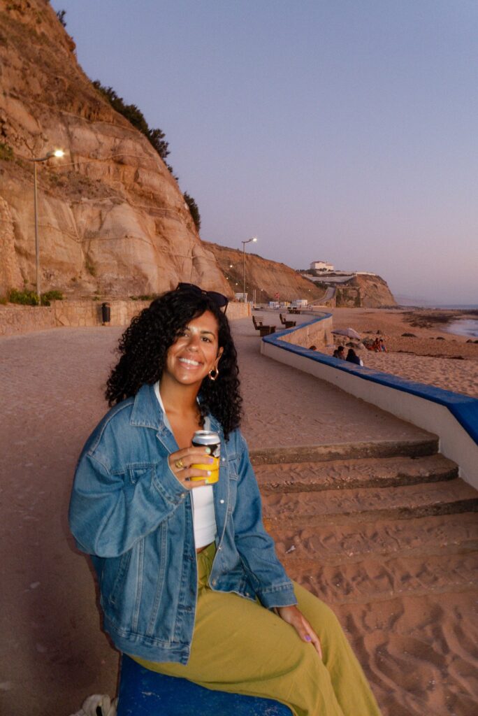 A woman with curly hair and a denim jacket smiles while sitting on a blue bench by the beach at sunset, holding a can—perhaps after exploring the Best Restaurants In Ericeira. Behind her are cliffs, a sandy path, and people enjoying the seaside.