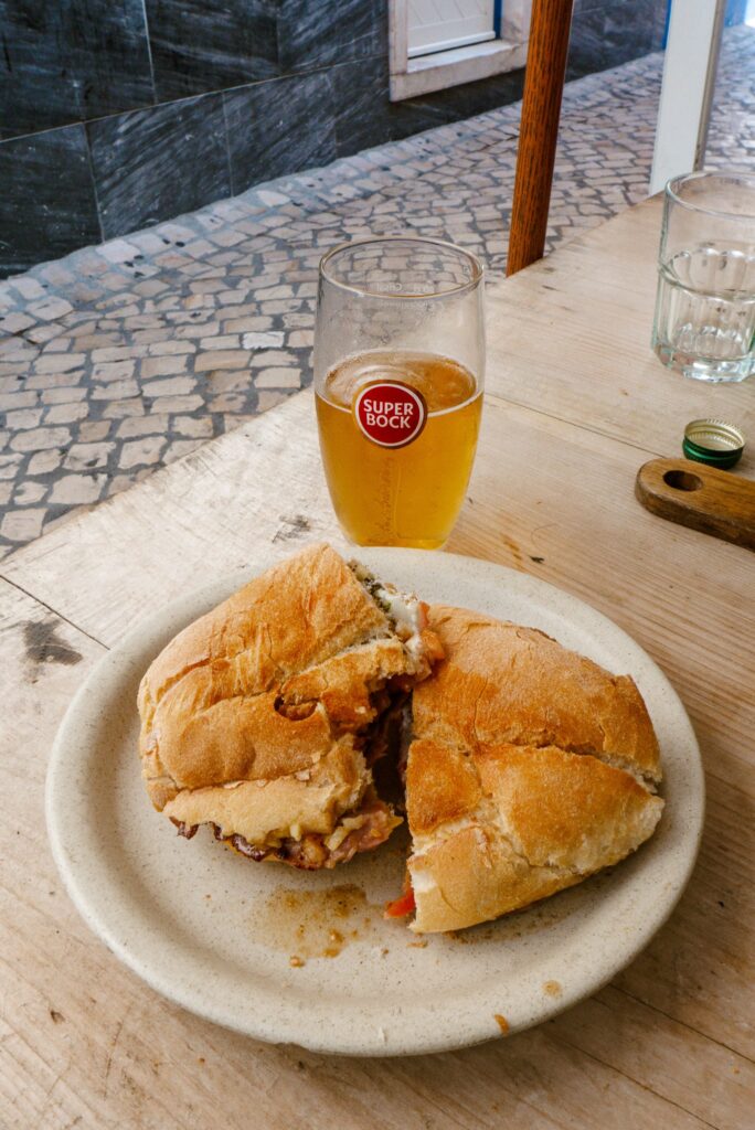 A plate with two halves of a sandwich sits on a wooden table next to a glass of Super Bock beer and an empty glass, capturing the relaxed vibe you’ll find at the Best Restaurants In Ericeira, with a cobblestone street visible in the background.