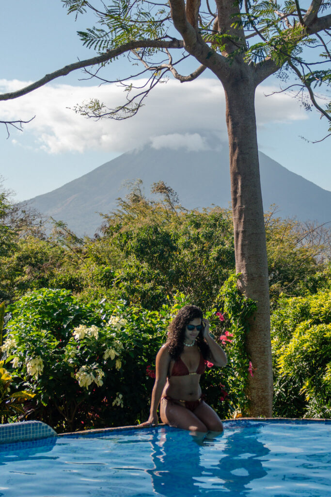 A woman in a red bikini sits at the edge of a swimming pool, surrounded by lush greenery, with a tall tree and a large volcano in the background—an idyllic stop on any 10-Day Nicaragua Itinerary under a partly cloudy sky.