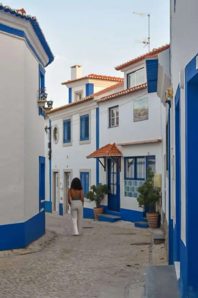 A woman with long hair walks down a narrow, cobbled street lined with white buildings trimmed in blue in sunlit Ericeira—capturing the charm of Portugal and inspiring thoughts of the Best Things To Do In Ericeira, Portugal.