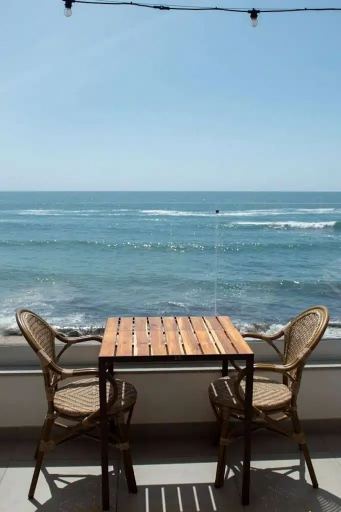 A wooden table with two wicker chairs is set on a balcony overlooking a calm, blue ocean under a clear sky—a tranquil escape and one of the Best Things To Do In Ericeira, Portugal. String lights hang above, gentle waves visible in the background.
