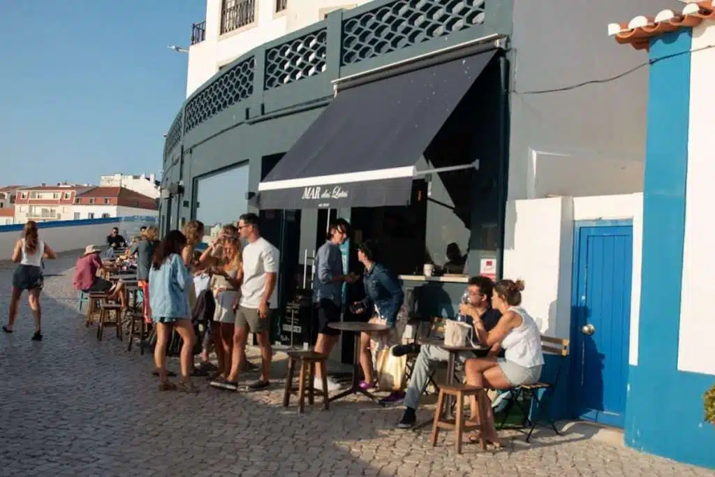 People sit and stand outside a small cafe with a black awning on a sunny day in Ericeira, Portugal, chatting and enjoying drinks. The cafe is on a cobblestone street with blue and white buildings—a perfect stop while exploring the Best Things To Do In Ericeira.