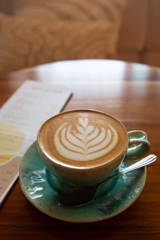 A cup of latte with leaf-shaped latte art sits on a turquoise saucer with a spoon, next to a blurred menu on a wooden table—perfect for planning the Best Things To Do In Ericeira, Portugal.