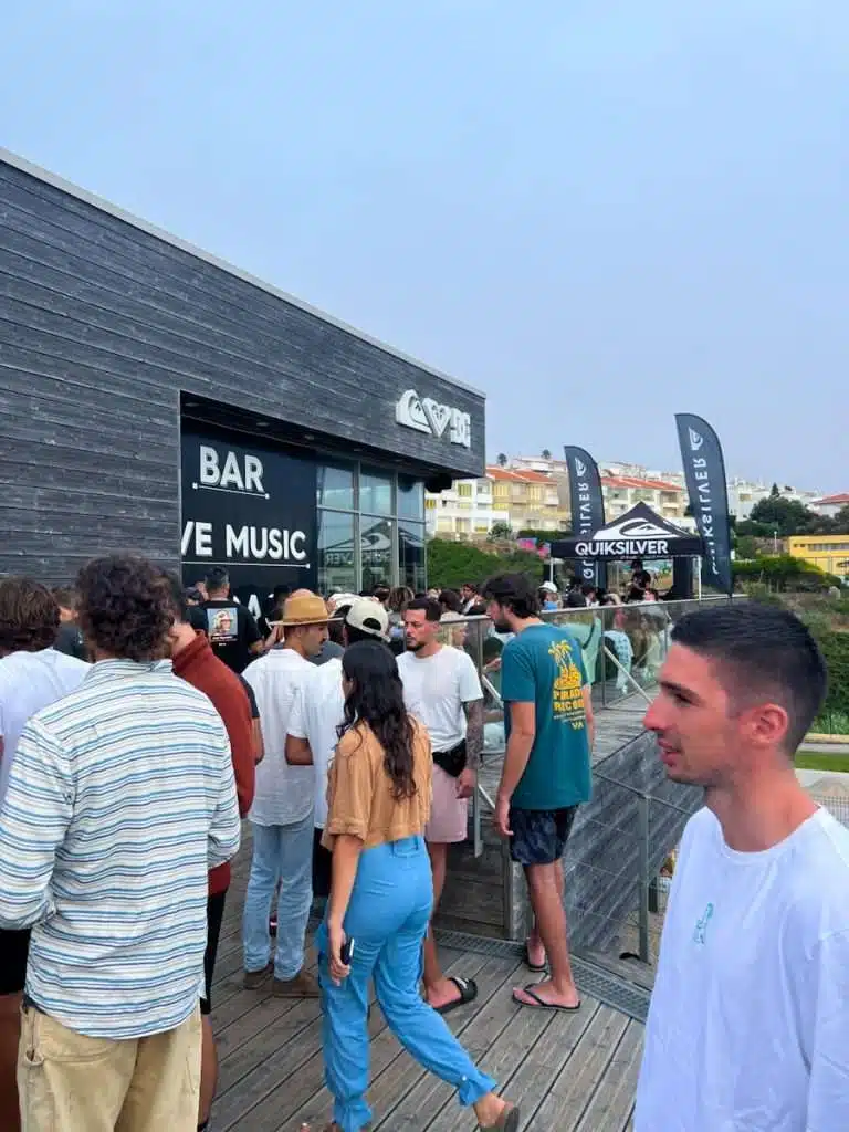 A crowd gathers outside a modern black building with “BAR” and “LIVE MUSIC” signs in Ericeira, Portugal. Some chat while others wait in line. Surf brand flags wave nearby—a true highlight among the Best Things To Do In Ericeira.