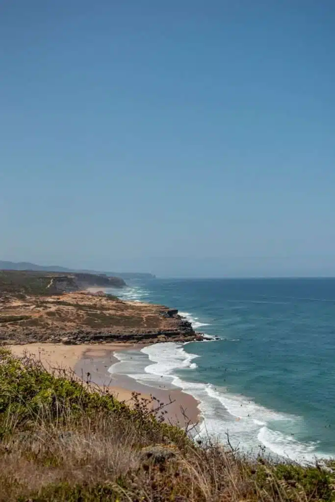 A scenic coastal view with sandy beaches, waves crashing on the shore, rocky cliffs, and green vegetation in the foreground under a clear blue sky—one of the Best Things To Do In Ericeira, Portugal.