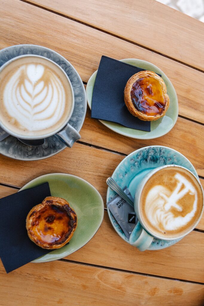 Two cups of latte art coffee and two Portuguese custard tarts (pastéis de nata) on colorful plates, arranged on a wooden table—an inviting scene at one of the Best Restaurants In Ericeira.
