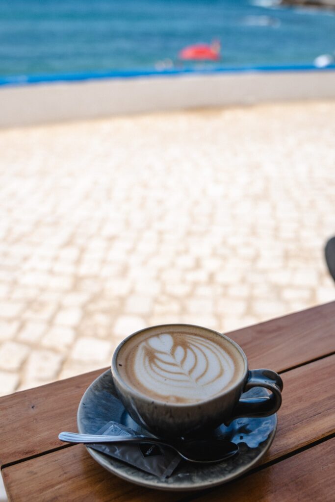 A cup of latte with foam art sits on a saucer with a spoon and napkin on a wooden table, overlooking a sunny seaside view with blue water in the background—an experience to savor at one of the Best Restaurants In Ericeira.