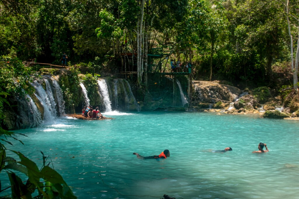People swim in a turquoise pool at the base of small waterfalls surrounded by lush greenery—a must-see on any Philippines island-hopping itinerary. Some float in life jackets, while others explore the scene from a bamboo raft framed by dense forest.