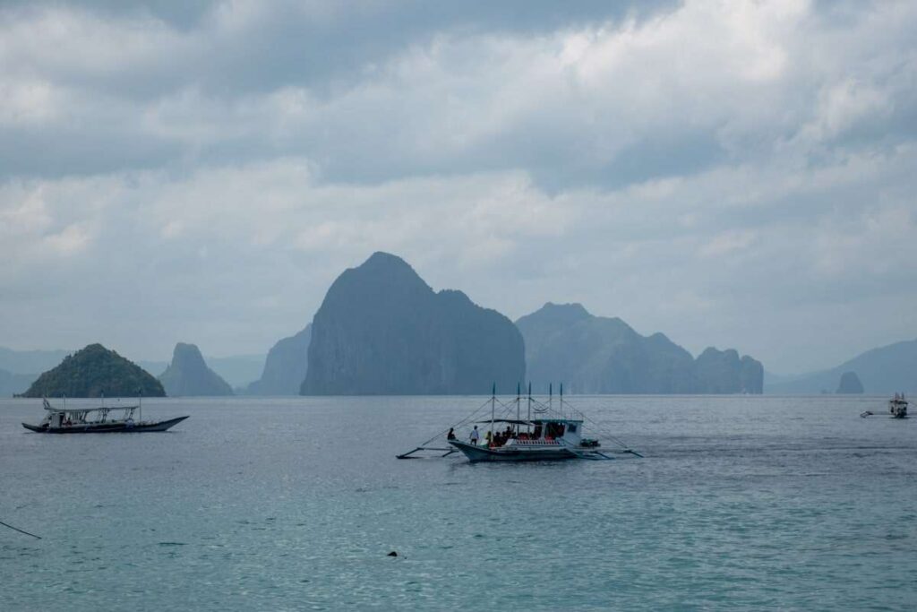 Traditional outrigger boats float on calm blue water with rocky islands and cloudy skies in the background—an idyllic setting perfect for any Philippines island-hopping itinerary. A few boats drift peacefully across this picturesque seascape.