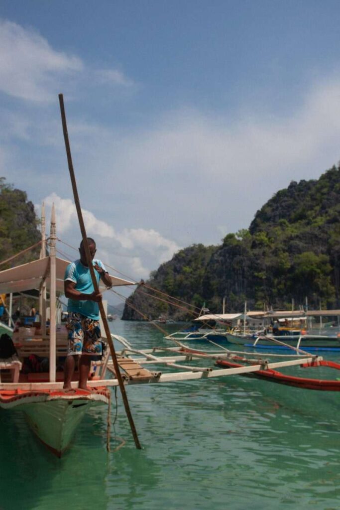 A man stands on a wooden boat holding a long pole in turquoise water—an iconic scene from a Philippines island-hopping itinerary, with more boats and lush, rocky hills in the background under a partly cloudy sky.