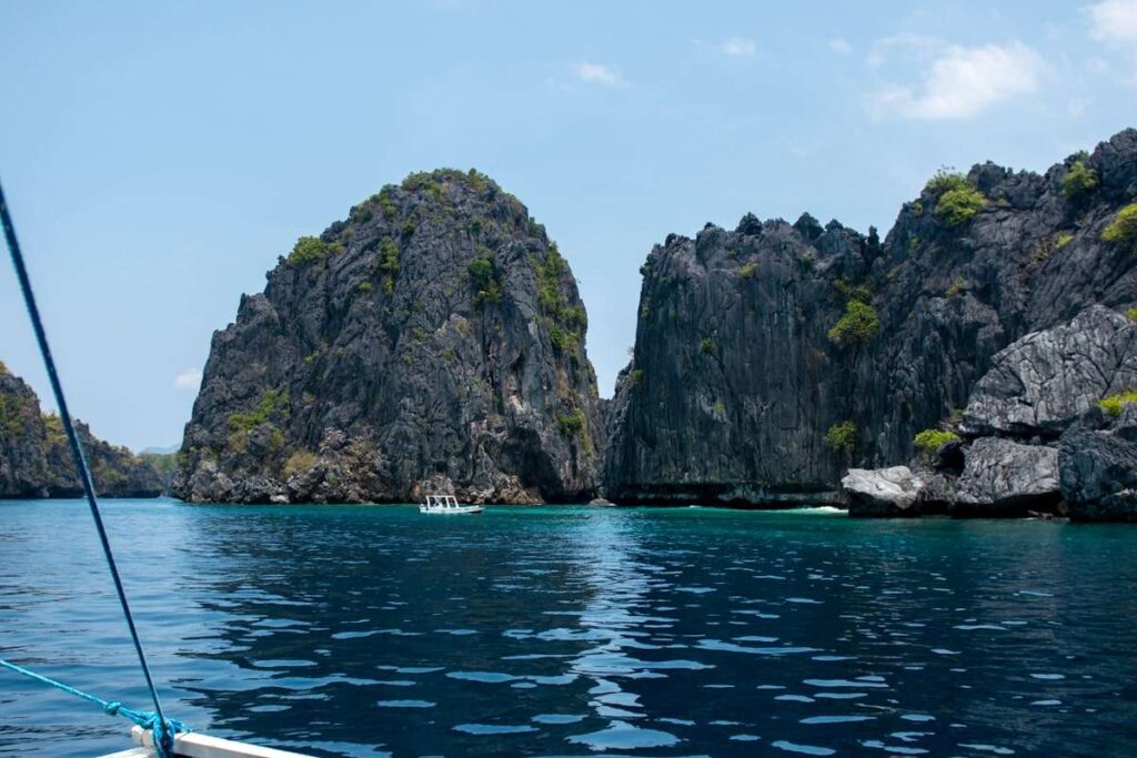A view from a boat of towering limestone cliffs covered with patches of greenery, surrounding clear blue water under a bright sky—an essential sight on any Philippines island-hopping itinerary. A small white boat is visible near the cliffs.
