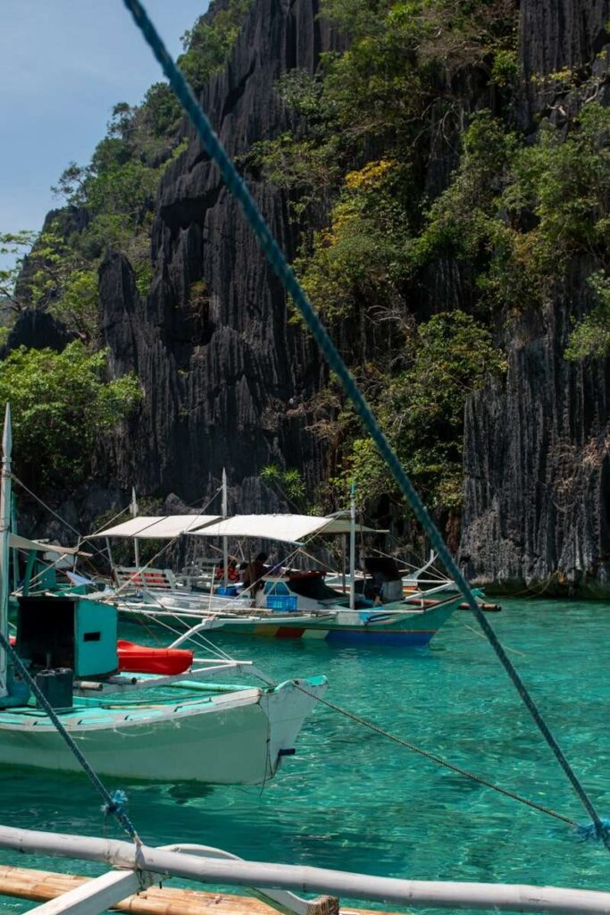 Traditional Filipino boats float on clear turquoise water near tall, rocky cliffs covered with lush vegetation—a stunning scene often featured in a Philippines island-hopping itinerary, all beneath a bright sky.