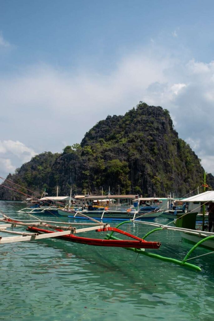 Traditional boats float on clear turquoise water near a lush, rocky island—an iconic scene on any Philippines island-hopping itinerary—complete with steep cliffs under a partly cloudy sky.