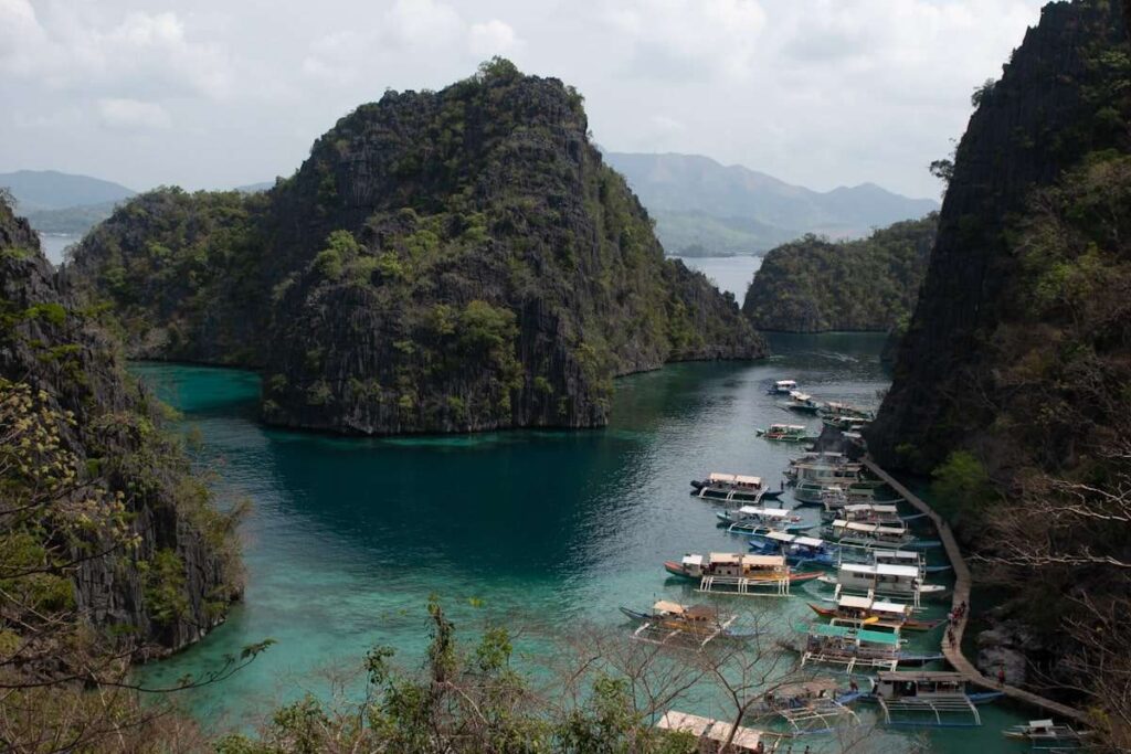A scenic lagoon surrounded by steep, rocky cliffs with lush greenery—a highlight on any Philippines island-hopping itinerary. Clear turquoise water fills the bay, where several boats are docked along a wooden pier on the right side of the image.