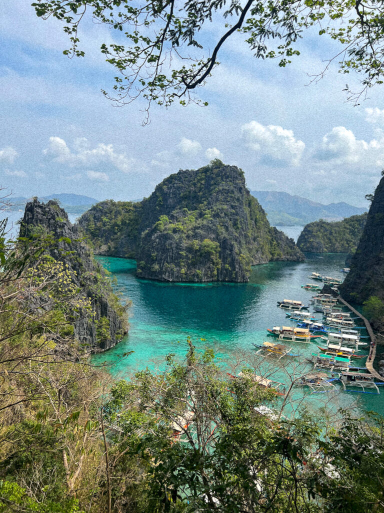 A scenic view of a tropical bay with turquoise water, dramatic limestone cliffs, and lush greenery—an idyllic stop on a Philippines island-hopping itinerary—where boats are docked along a wooden pier, framed by overhanging branches and foliage.