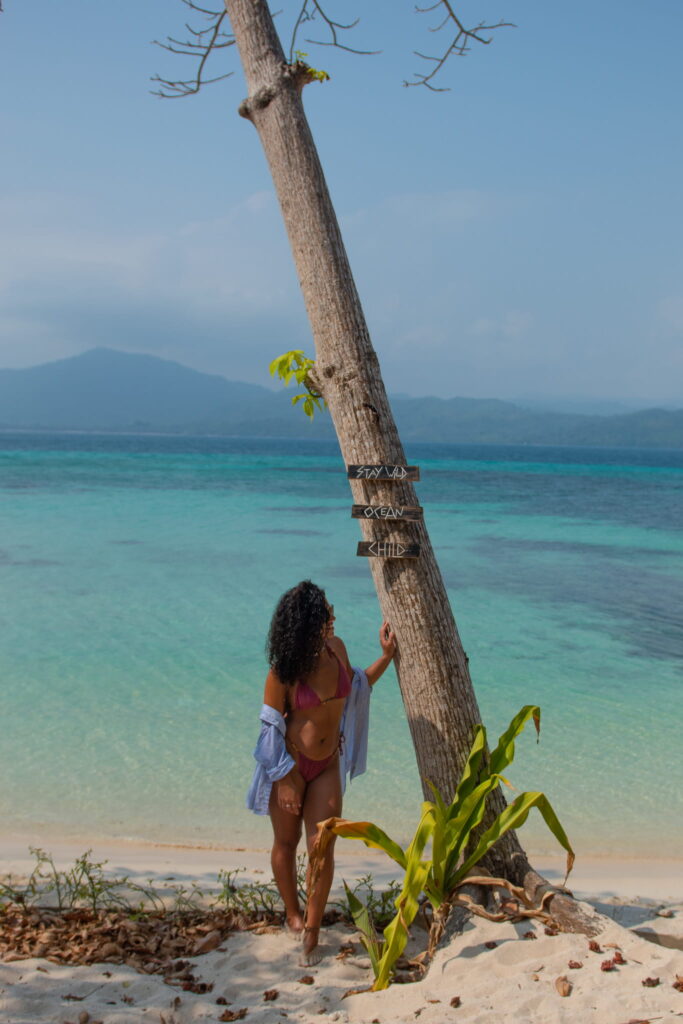 A woman in a bikini and open shirt stands on a sandy beach, touching a leaning palm tree with wooden signs—an inviting scene for any Philippines island-hopping itinerary, with clear turquoise water and distant mountains under a blue sky.