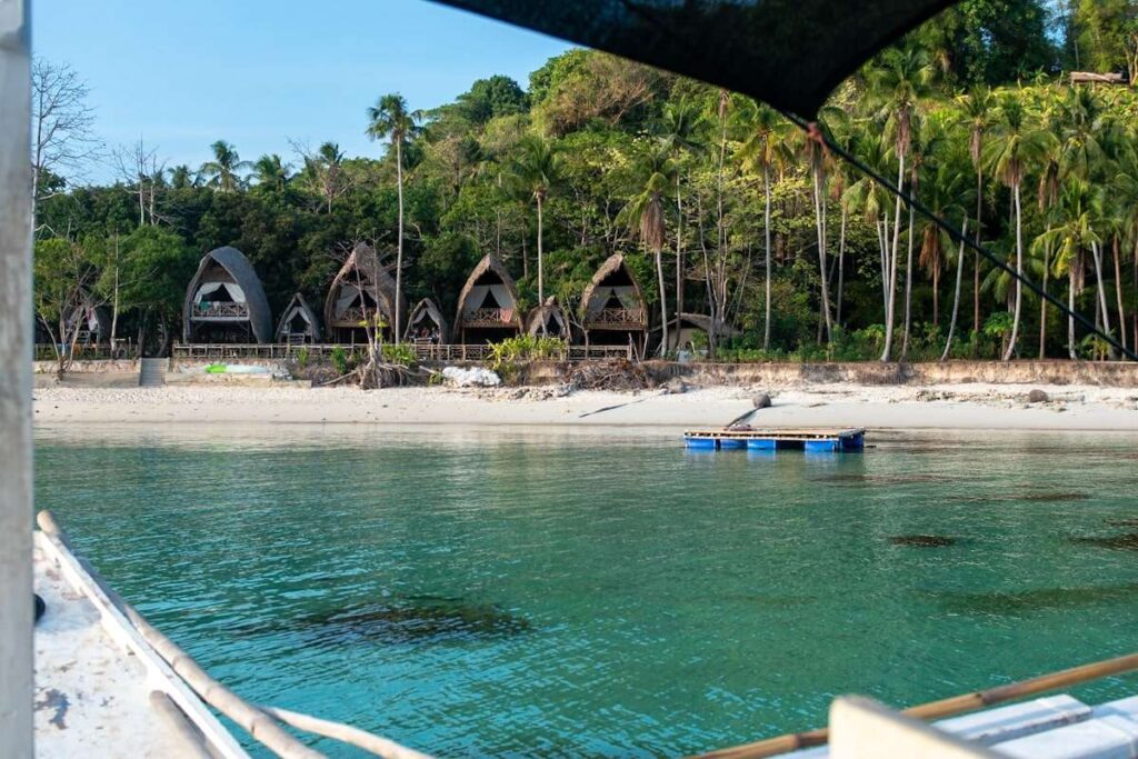 A tranquil beach scene with clear turquoise water, a sandy shore, and unique, arch-shaped wooden huts nestled among lush palm trees—an idyllic stop on your Philippines island-hopping itinerary. Part of a boat is visible in the foreground.