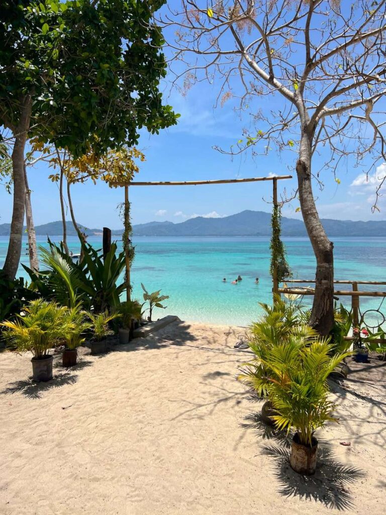 A sandy beach with potted plants and trees frames a bamboo archway, looking out onto clear turquoise water—an idyllic stop on any Philippines island-hopping itinerary, where people swim beneath a blue sky with mountains in the distance.