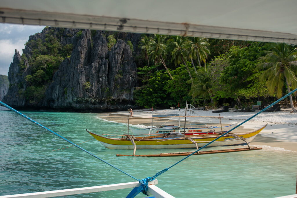 A yellow outrigger boat floats on turquoise water near a white sandy beach, perfect for any Philippines island-hopping itinerary, surrounded by lush green palm trees and tall limestone cliffs under a partly cloudy sky.