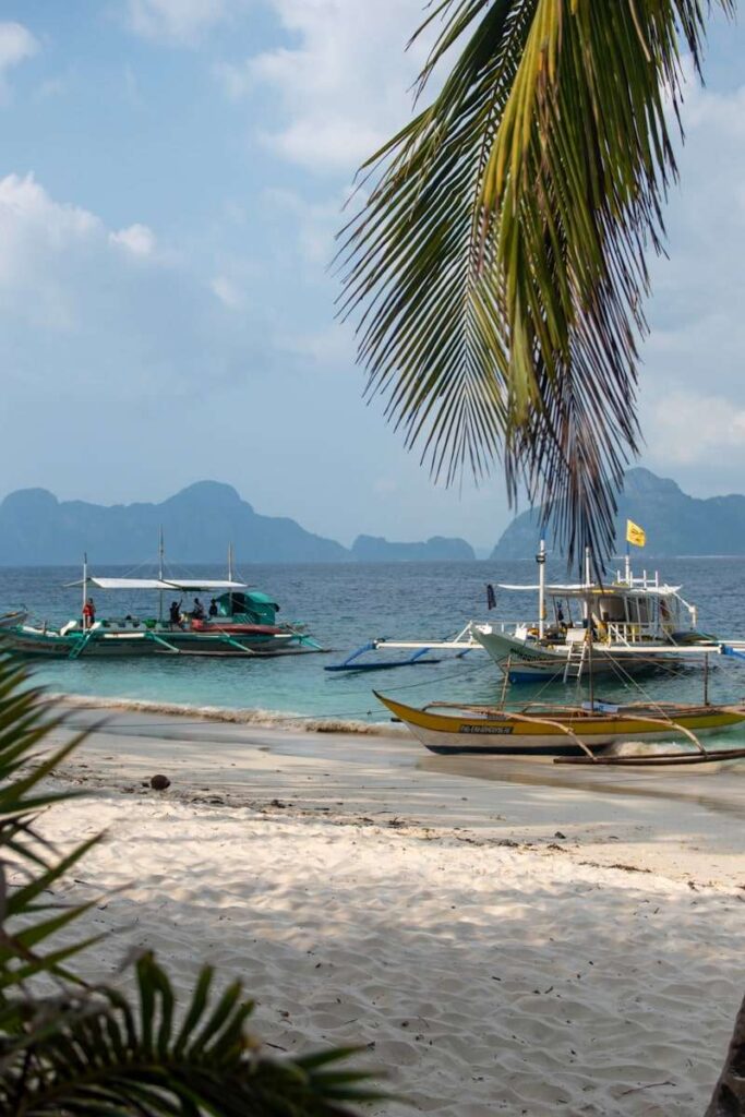 Three traditional outrigger boats float near the shore of a sandy beach, palm leaves framing this classic view—an inviting stop on any Philippines island-hopping itinerary, with blue water and distant limestone islands under a partly cloudy sky.