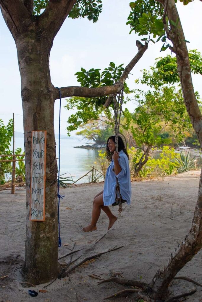A woman in a blue shirt sits on a rope swing beneath the trees on a sandy beach, embracing the Isla Experience. A sign on the tree reads, "PICK YOUR SELF, IT'S NOT FRAGILE," with water and trees in the background.