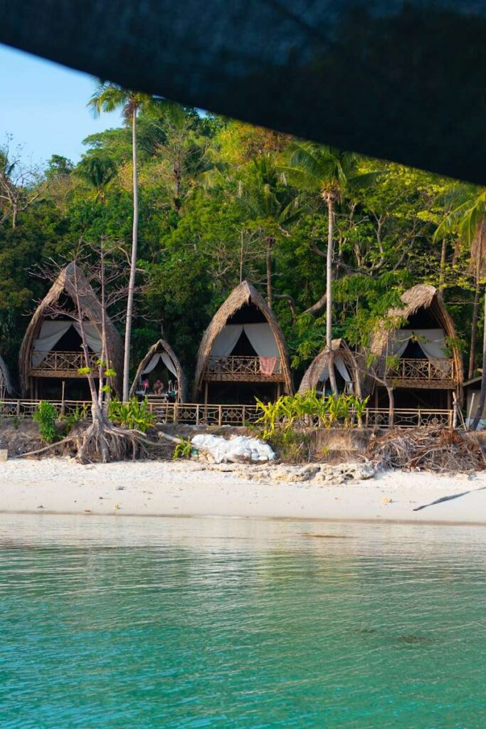 Three thatched-roof huts on a sandy beach offer an authentic Isla Experience, surrounded by lush green trees, with clear turquoise water in the foreground and a dark fabric partially covering the top of the image.