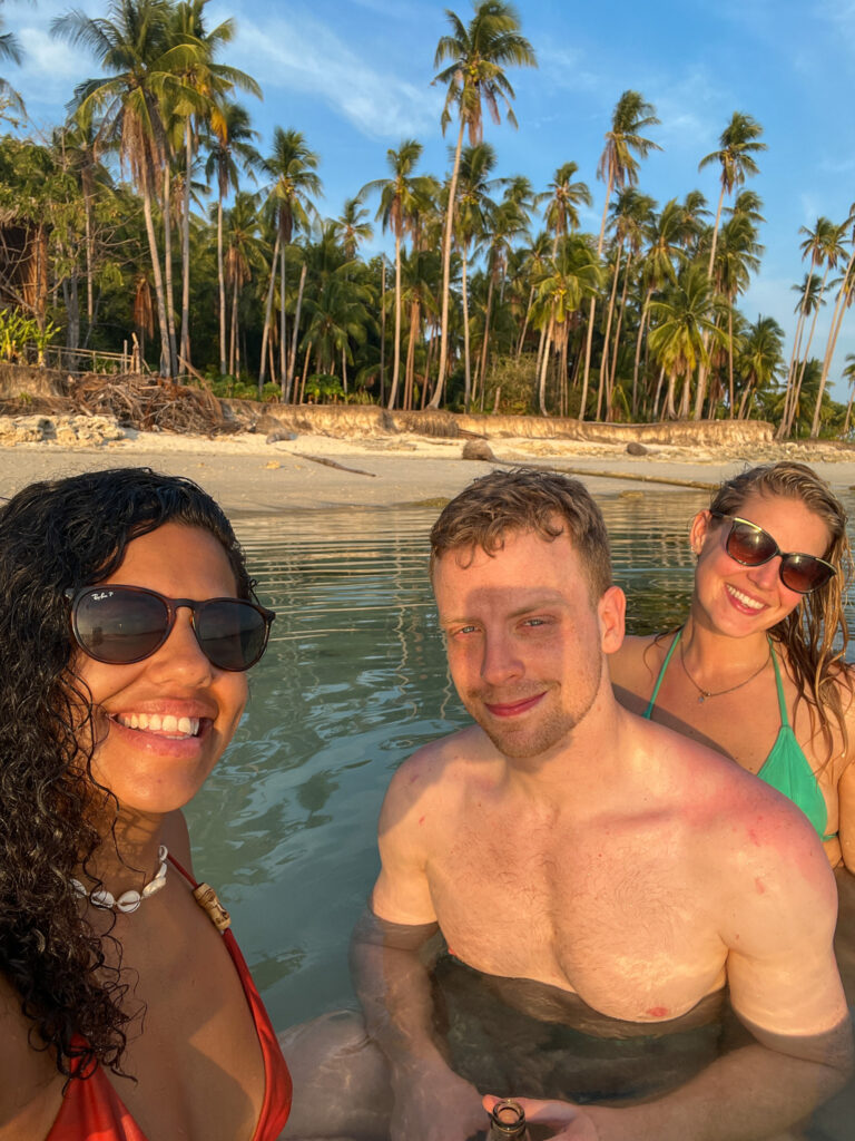 Three people smiling and posing for a photo while standing in shallow water at a tropical beach, enjoying Isla - The Island Experience, with palm trees and a sandy shore in the background under a clear blue sky.