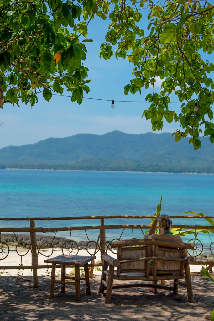 A person sits on a bamboo bench under leafy trees, gazing out at clear turquoise water and distant green mountains—immersed in Isla - The Island Experience on this sunny beach.