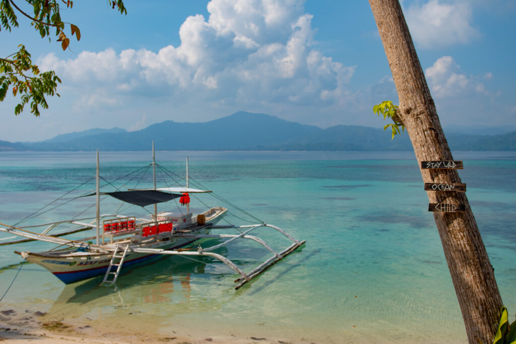 A traditional outrigger boat is anchored in clear turquoise water near a sandy beach. A tree with signs reading "STAY WILD," "OCEAN," and "CHILD" welcomes you to Isla - The Island Experience, with mountains and clouds across the bay.