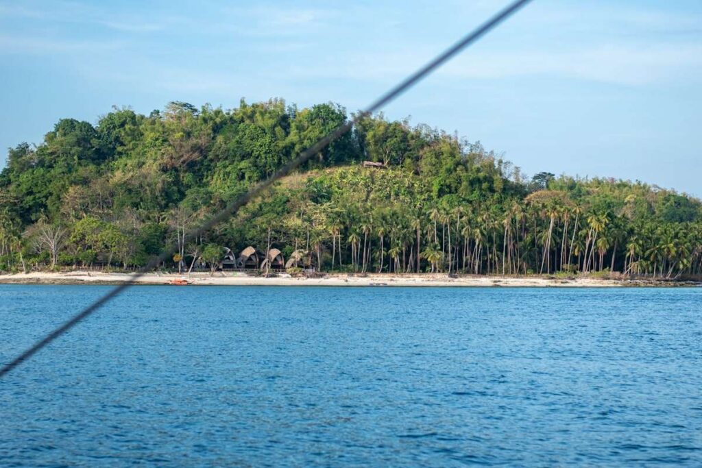A lush green island with palm trees, rustic huts along the sandy shore, and blue ocean water in the foreground offers a true Isla Experience. A diagonal wire or rope crosses the image beneath a clear, bright sky.