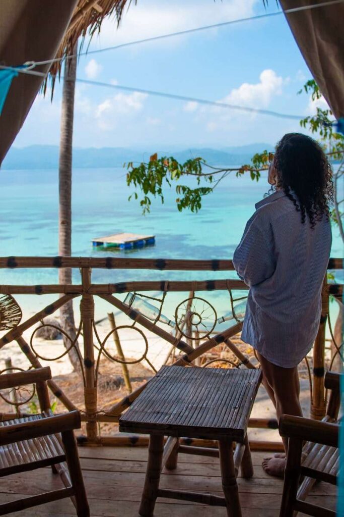 A person in a long shirt stands on a bamboo balcony, enjoying the Isla Experience—overlooking clear turquoise water, a wooden dock, palm trees, and distant mountains beneath a blue sky.